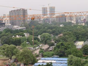 Urban landscape with green trees, an unpaved road, construction crane, and tall buildings under an overcast sky.