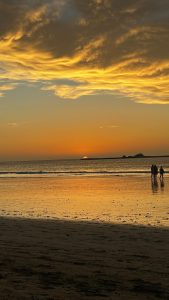 Sunset over a calm beach with glowing clouds, reflective water, and people walking along the shore.
