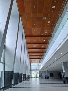 Low-angle wide shot of a spacious, modern building lobby with high ceilings featuring warm wooden paneling, tall glass window walls on the left, and a multi-level mezzanine corridor with light grey tiled flooring on the right.