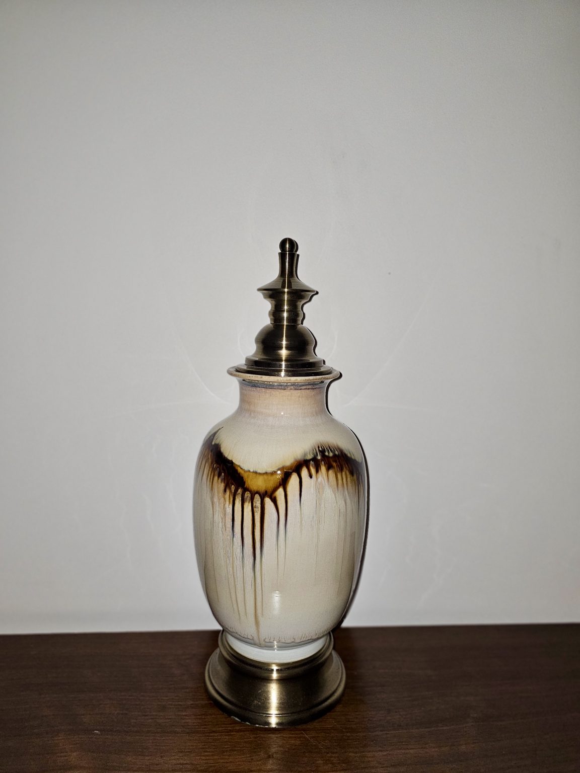 A decorative ceramic bottle with a metallic lid stands on a wooden surface. Its pale body has flowing brown glaze marks, while the dark metal top and base add contrast. Captured in Chalappuram, Kozhikode, Kerala.