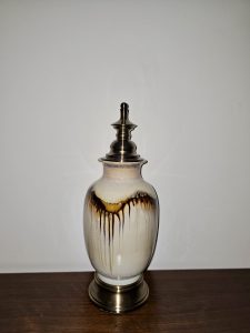 A decorative ceramic bottle with a metallic lid stands on a wooden surface. Its pale body has flowing brown glaze marks, while the dark metal top and base add contrast. Captured in Chalappuram, Kozhikode, Kerala.