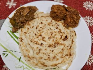 A stack of flaky Kerala-style parottas is served with portions of chicken and squid curry on a white plate. The layered texture of the parotta and the rich colors of the curries create an inviting food photograph. Captured in Koolimadu, Kozhikode, Kerala.