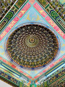 A richly decorated Chinese temple ceiling features a large circular dome surrounded by colorful geometric patterns, golden dragons, birds, and intricate floral designs. The detailed artwork creates a striking radial composition. Captured at Thean Hou Temple, Kuala Lumpur, Malaysia.