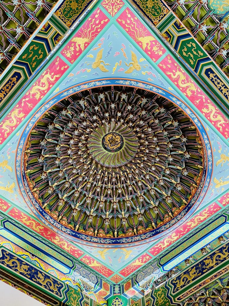 A richly decorated Chinese temple ceiling features a large circular dome surrounded by colorful geometric patterns, golden dragons, birds, and intricate floral designs. The detailed artwork creates a striking radial composition. Captured at Thean Hou Temple, Kuala Lumpur, Malaysia.