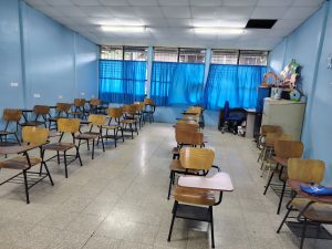 
A classroom with wooden desks, blue walls, and large windows letting in natural light.