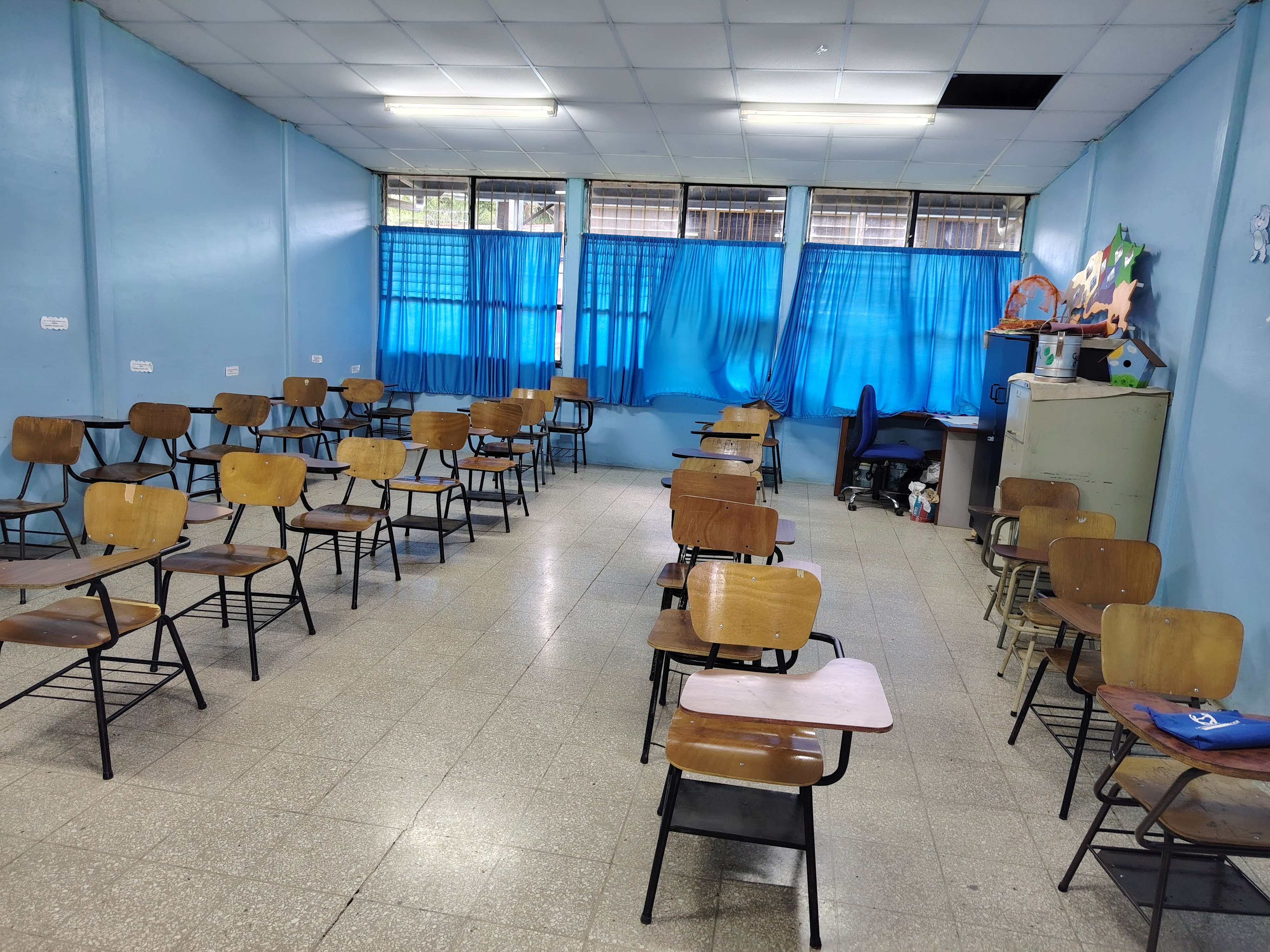 A classroom with wooden desks, blue walls, and large windows letting in natural light.