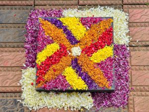 A colorful square Pookkalam made with flower petals in shades of orange, yellow, red, white, pink, and purple. The floral design is placed on a decorative tiled floor, creating a bright and festive Onam display. Location: Perumanna, Kozhikode, Kerala.