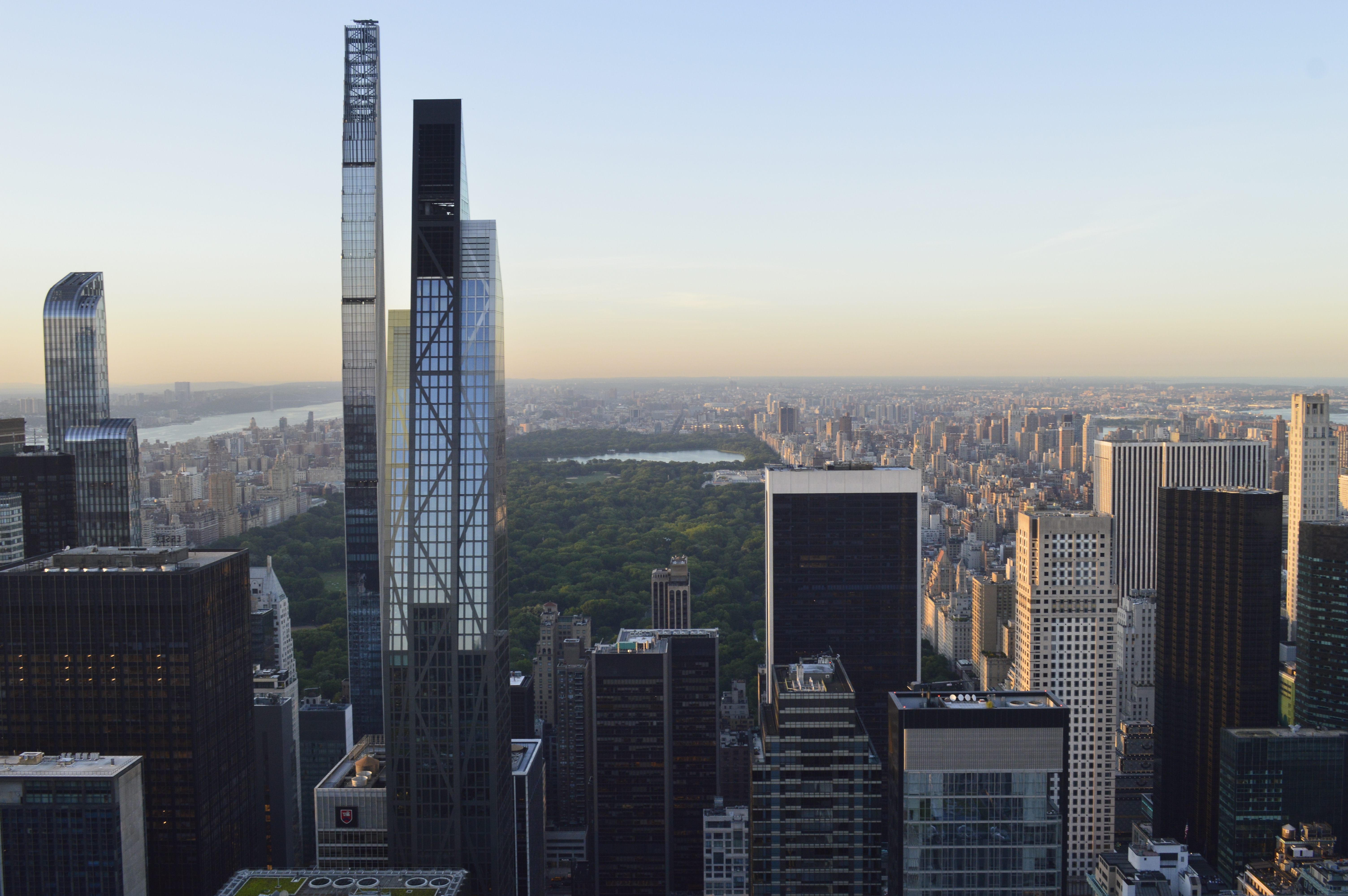 A panoramic view of a large city skyline with tall modern skyscrapers overlooking a sprawling green urban park under a clear evening sky