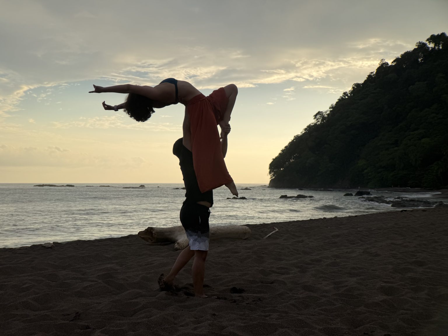 Two people in an acrobatic balance on a beach at sunset, one arched back high above the other.