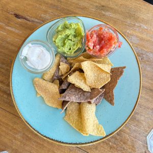 A round turquoise plate rests on a wooden table, filled with a variety of tortilla chips in shades of yellow and brown.
