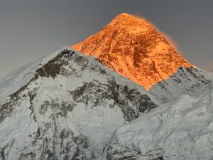 Mount Everest while sunrise. The highest peak is illuminated with a warm, orange glow. Surrounding mountains appear stark and dramatic, composed of harsh rock and ice formations.