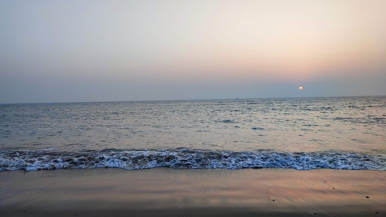 Saint Martin’s sea beach at sunset, with gentle waves rolling onto the sandy shore, a calm blue sea, and the sun setting near the horizon.