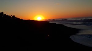 A bright orange sunset over a dark beach, with ocean waves on the right and silhouettes of palm trees and people along the shore.