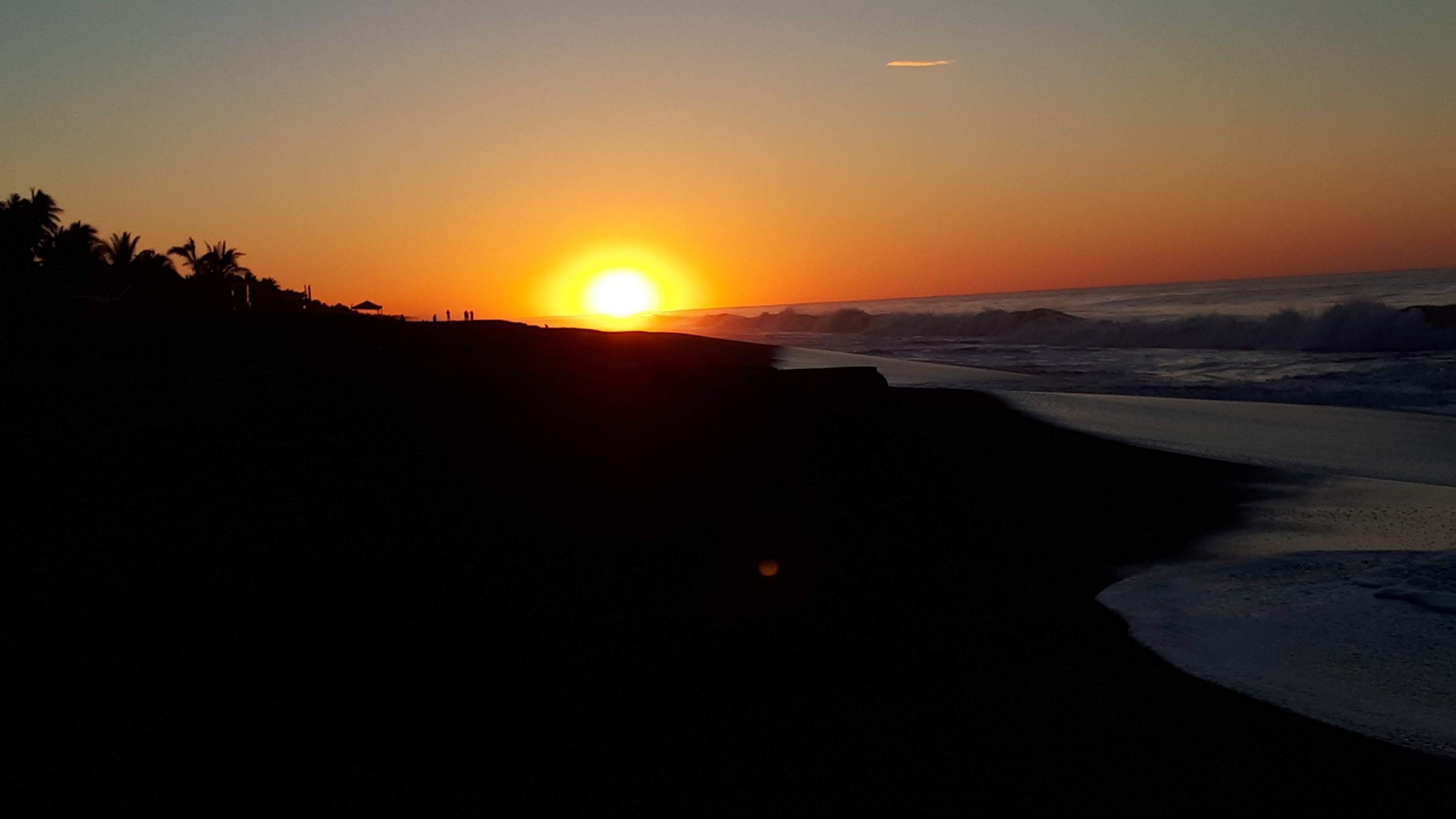 A bright orange sunset over a dark beach, with ocean waves on the right and silhouettes of palm trees and people along the shore.
