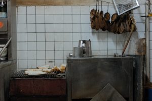 A traditional coffee shop kitchen setup featuring bread toasting over a charcoal grill next to metal tea kettles, with cloth-strainers hanging against a white-tiled wall.