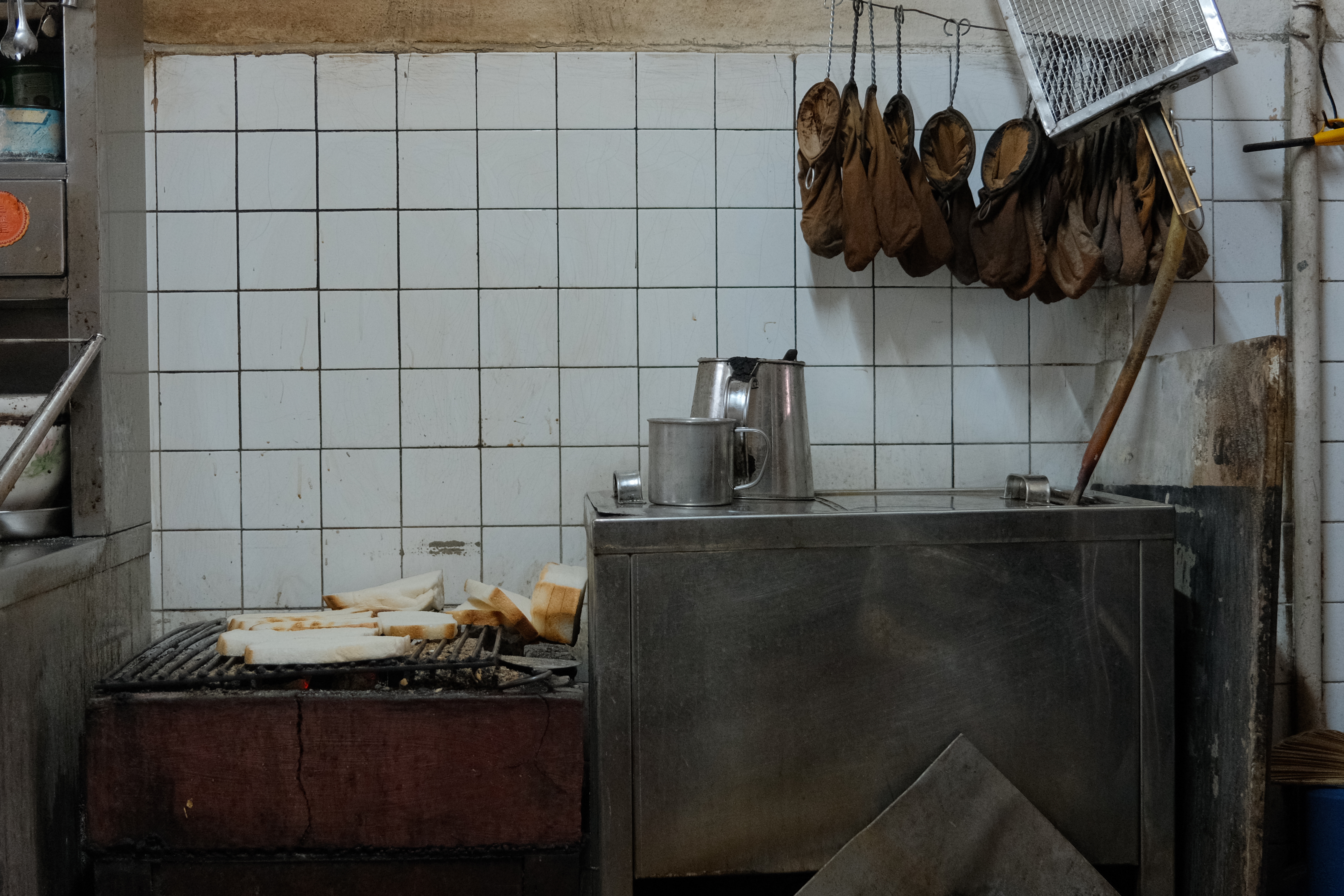 A traditional coffee shop kitchen setup featuring bread toasting over a charcoal grill next to metal tea kettles, with cloth-strainers hanging against a white-tiled wall.