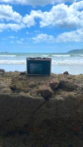 
A small digital camera is positioned on a textured stone surface, with the ocean in the background.