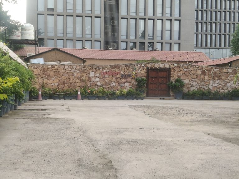 A rustic wooden entrance in a stone wall, surrounded by potted green plants, with a modern glass building in the background.