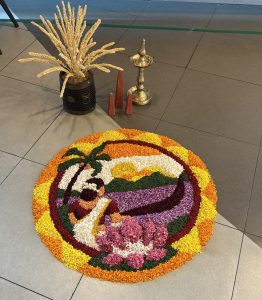 A colorful floral rangoli with a traditional figure, lotus flowers, and palm trees, beside a decorative pot and metal lamp.