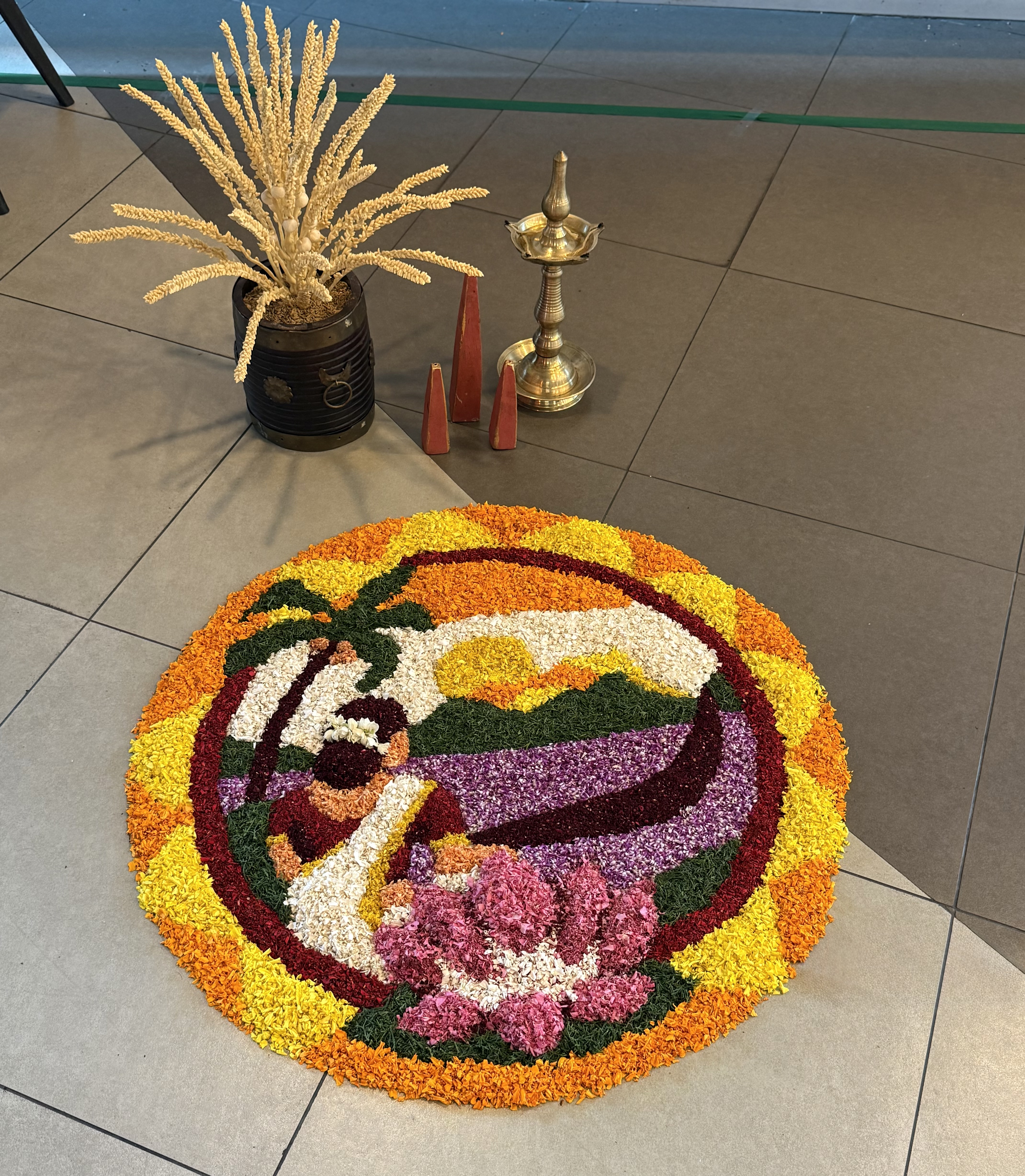 A colorful floral rangoli with a traditional figure, lotus flowers, and palm trees, beside a decorative pot and metal lamp.