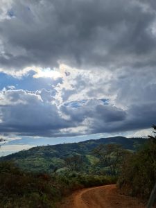 Dark storm clouds gather above rolling green hills, while a winding dirt road leads through the landscape beneath a brief opening of blue sky
