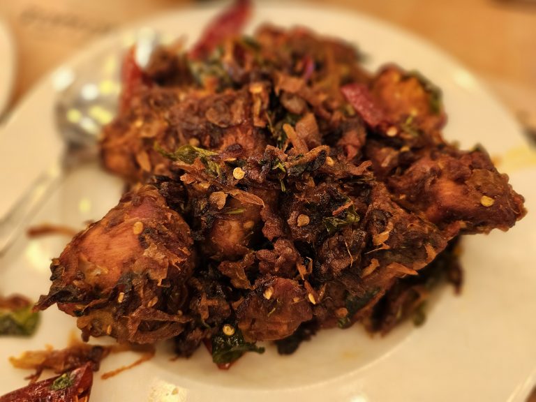 A close-up photograph of crispy fried chicken pieces (Chicken Chukka) served on a white plate. The dish is covered with fried onions, curry leaves, and spices, creating a rich, rustic appearance. The shallow focus draws attention to the food. Location: Perumanna, Kozhikode, Kerala.