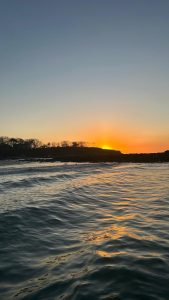 A peaceful ocean sunset with gentle waves, a glowing sky, and a distant hillside silhouette.