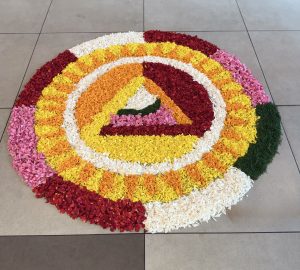 A colorful circular floral design with red, pink, yellow, white, and orange petals surrounding a green center.