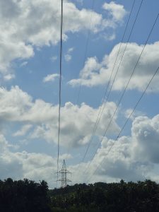 A blue sky filled with large white clouds above a green coconut-covered landscape. High-voltage power lines and a transmission tower add strong leading lines and an interesting contrast to the natural scenery. Location: Pantheerankavu, Kozhikode, Kerala.