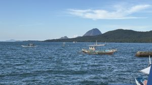 Colorful outrigger boats float on rippling blue coastal waters, with forested islands and a pointed mountain beneath a clear, sunny sky.