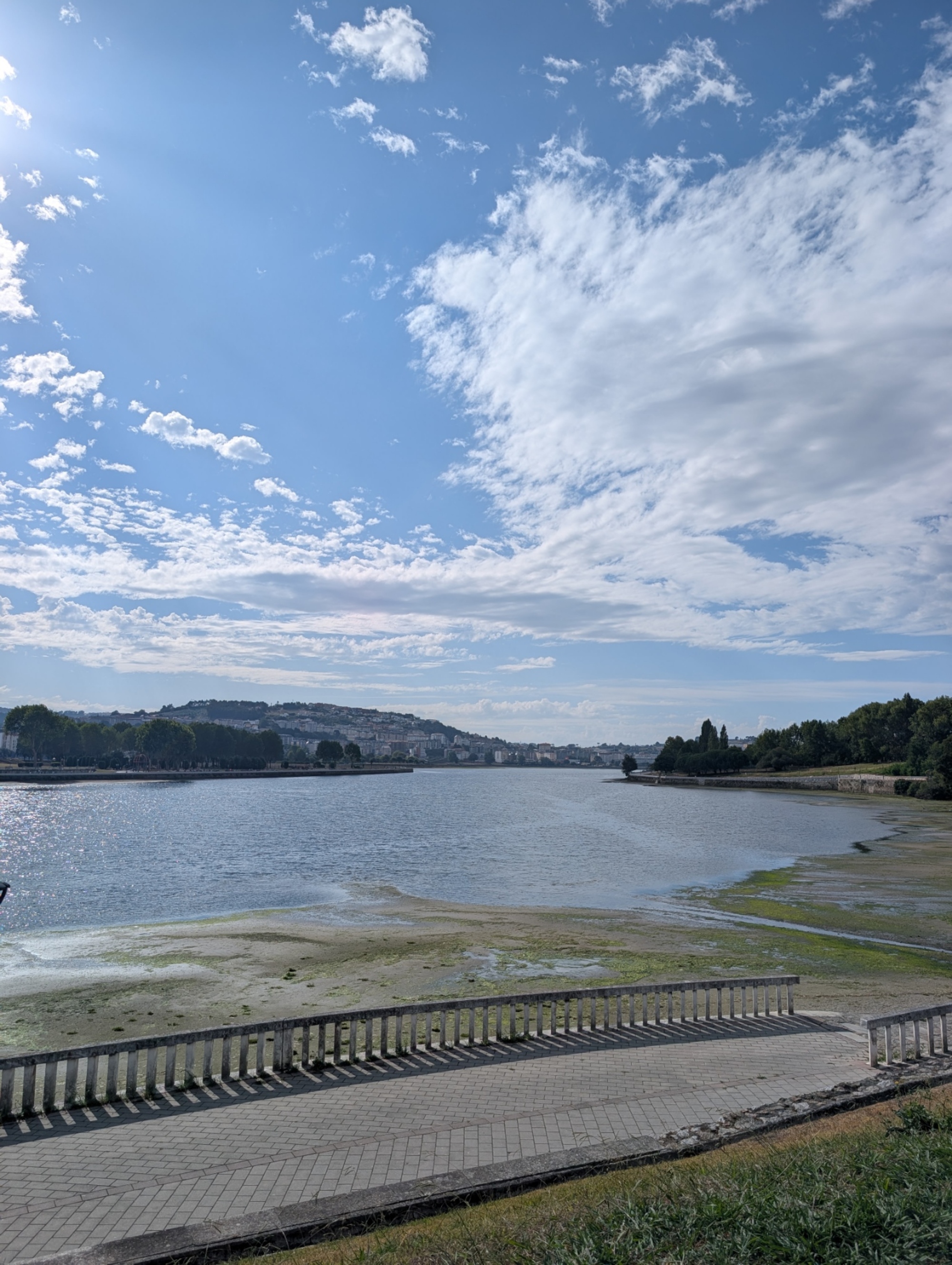 Wide scenic view of a coastal estuary promenade with low tide mudflats, surrounding green hills, and light clouds scattered across a bright blue sky.