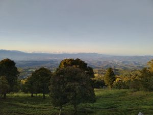 A panoramic landscape view captures a lush, green valley surrounded by rolling hills and distant mountains under a clear sky. 