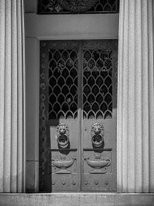 Doors to a mausoleum in a cemetery. The doors are ornate with lions holding rings in their mouths as the door handles. Large columns are to either side.
