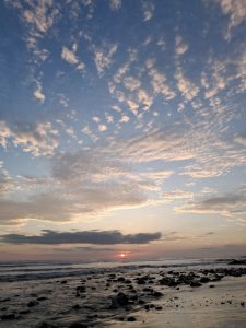 Sunset beach with gentle waves, dark rocks, and wet sand reflecting the fading light.