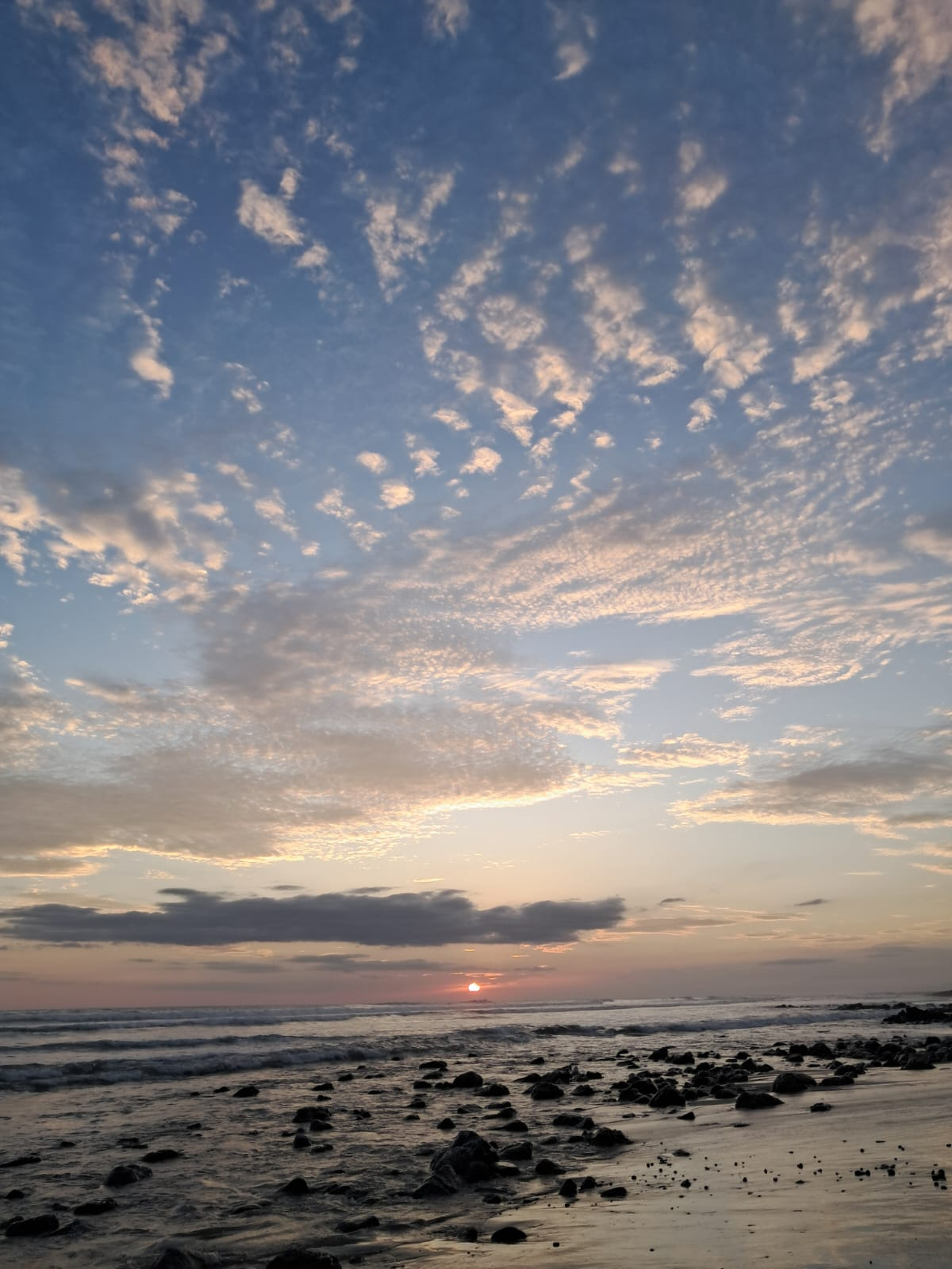 Sunset beach with gentle waves, dark rocks, and wet sand reflecting the fading light.