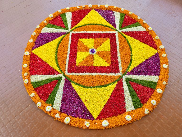 A large and colorful Pookkalam arranged in a circular geometric pattern with red, yellow, orange, purple, white, and green flower petals. The wide composition shows the complete design and its decorative details. Location: Perumanna, Kozhikode, Kerala.