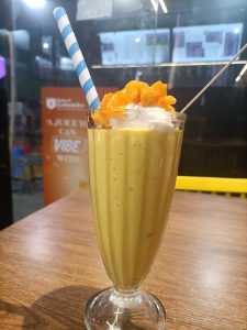 Creamy mango juice topped with whipped cream and mango pieces in a glass with a striped straw.
