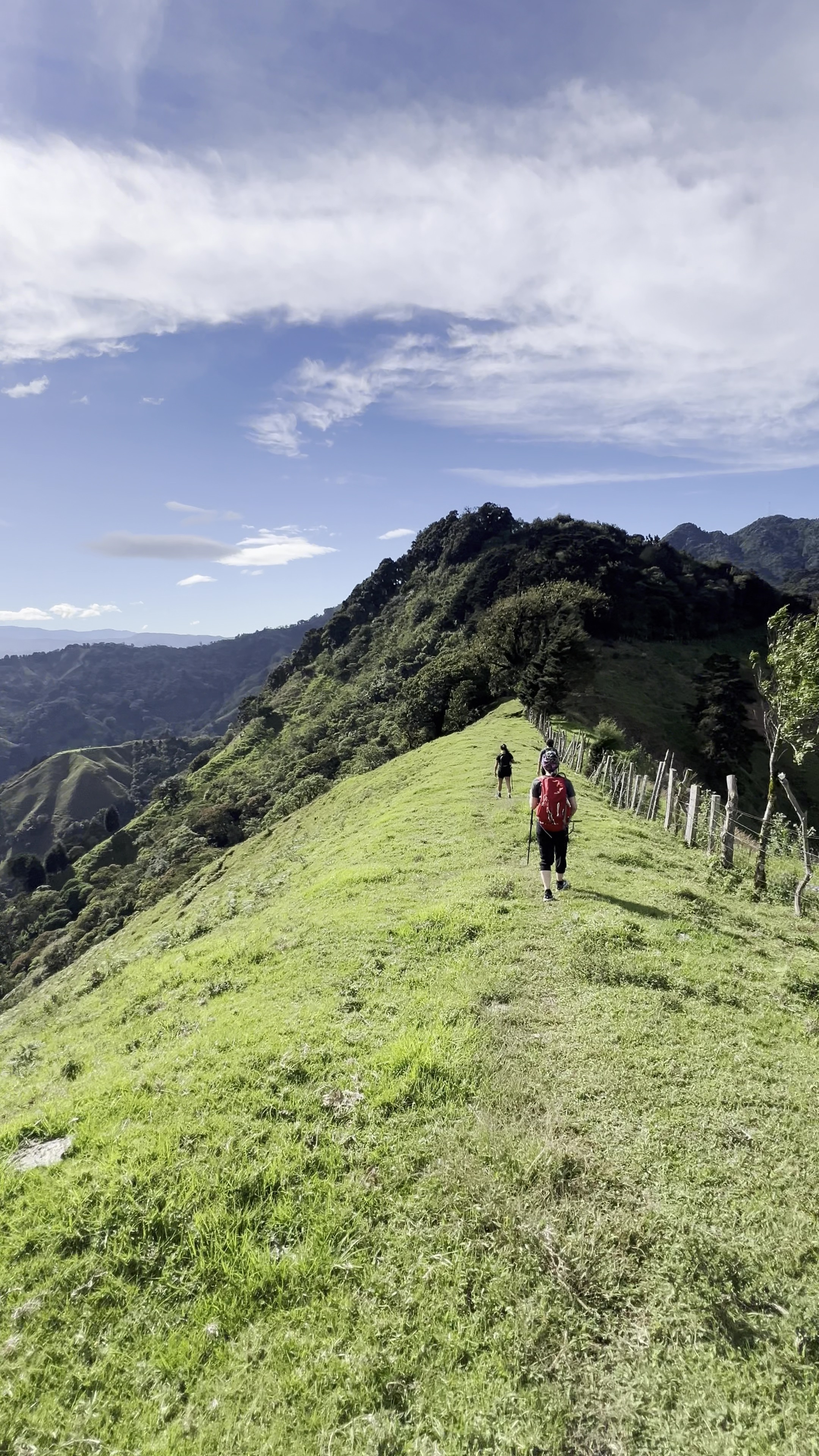 Two hikers walk along a grassy hillside trail with mountain views under a bright blue sky.