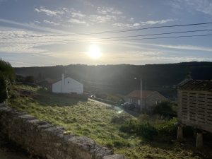 
Rural Galician landscape with traditional houses, green fields, a small road, and distant hills at sunset.