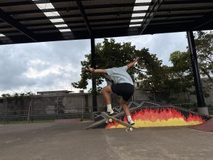 A young person is performing a skateboard trick in a park setting. They are captured mid-air, having just jumped off a ramp. The skateboarder is wearing a light blue shirt and black shorts, with their legs bent and arms extended for balance. Below them, there's a colorful ramp designed with a fiery gradient of red, orange, and yellow.
