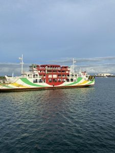 A large white passenger ferry with prominent red, green, and yellow painted accents floating on blue water under a partly cloudy sky.