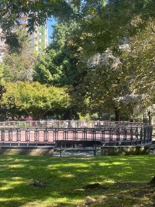 A circular wooden platform is surrounded by a black metal railing, set in a park area that features lush green grass and a variety of trees