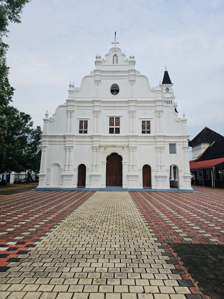 A symmetrical frontal view of an ornate white historic church, with its decorative façade, central doorway, tower, and patterned courtyard clearly visible. Location: Ramapuram, Kottayam, Kerala.