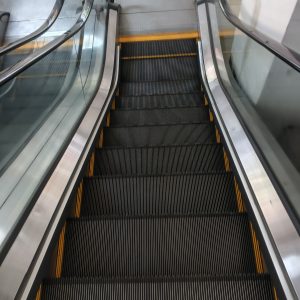 Downward escalator with black ridged steps, yellow edge lines, silver sides, and glass panels.