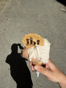 A hand holding a close-up of an ice cream cone with two scoops: one of stracciatella, which is beginning to melt, and another of Ferrero Rocher. A wafer-like cookie with the "La ibi Helados" logo is topped with the ice cream.