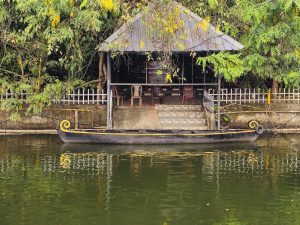 A traditional Kerala boat rests along the calm water beside a small riverside pavilion, surrounded by lush greenery and yellow flowers. The reflections create a peaceful scene. Location: Ayamkudy, Kottayam, Kerala.
