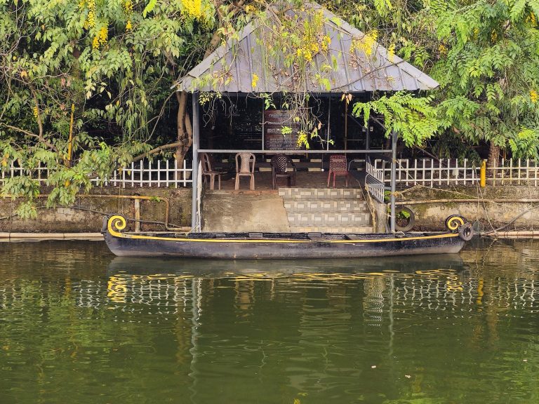 A traditional Kerala boat rests along the calm water beside a small riverside pavilion, surrounded by lush greenery and yellow flowers. The reflections create a peaceful scene. Location: Ayamkudy, Kottayam, Kerala.