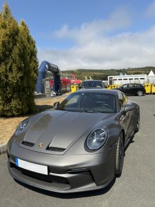 Gray metallic Porsche sports car parked on an outdoor racetrack, with a blue event arch, other cars, tents, and green hills under a blue sky.