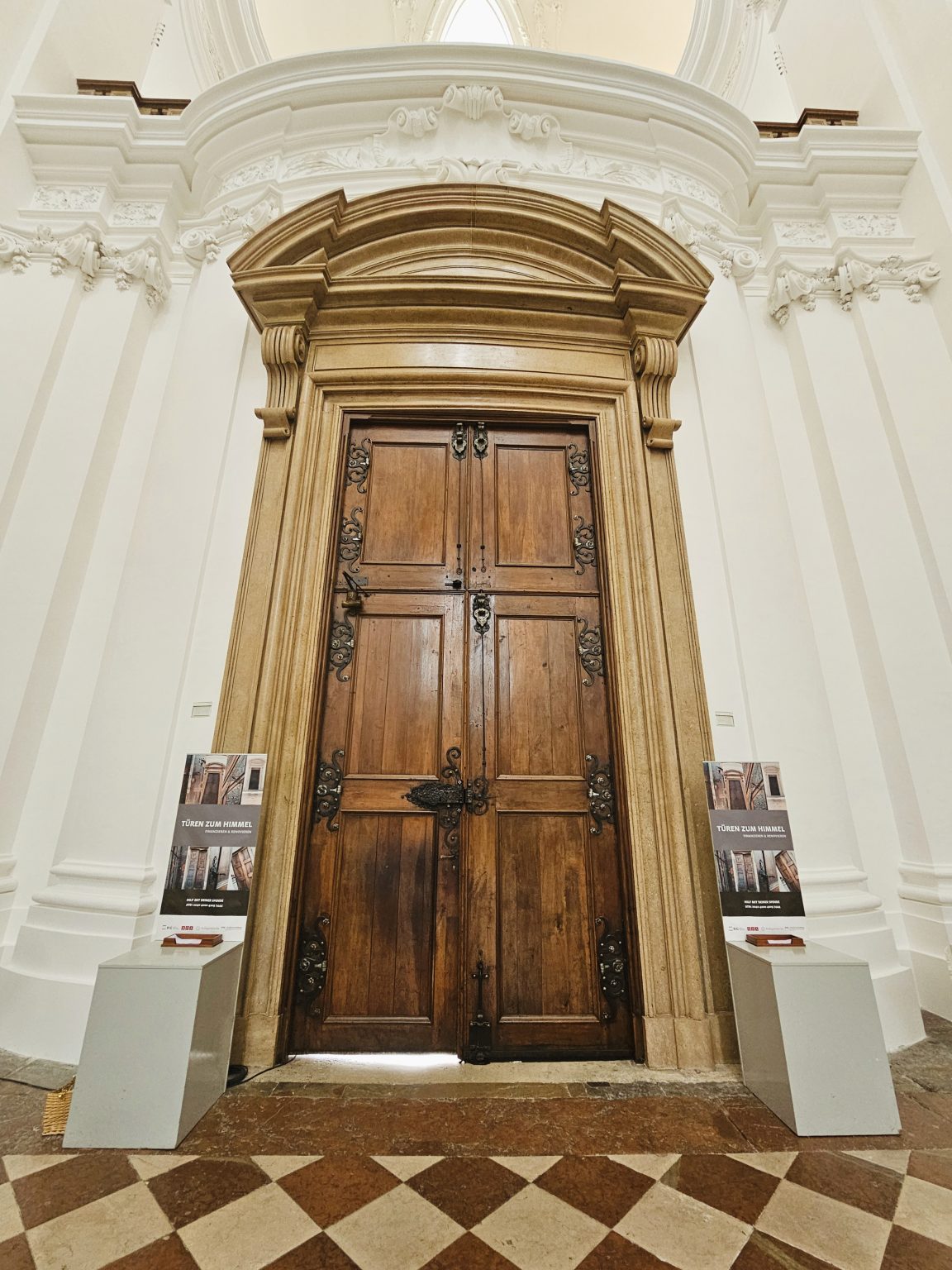 An ornate historic wooden doorway stands inside a richly decorated white Baroque interior, with intricate architectural details and a patterned stone floor. Location: Salzburg, Austria.