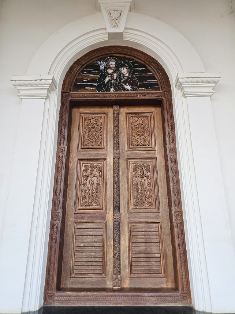A beautifully carved wooden doorway featuring a religious artwork panel above it. The symmetrical design, detailed carvings, and white architectural frame create an elegant composition. Location: Ramapuram, Kottayam, Kerala.